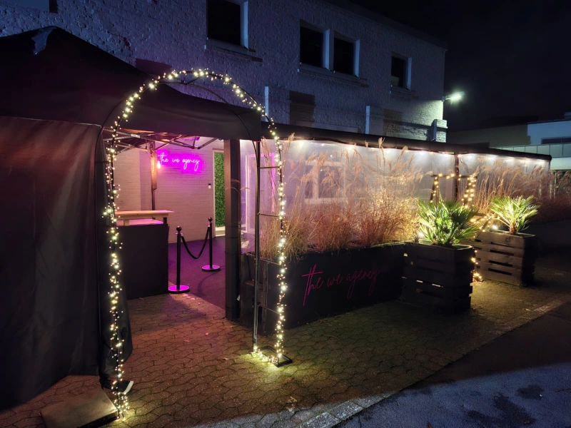 Beleuchteter Eingangsbereich der The We Agency Eventlocation bei Nacht mit Lichterketten-Bogen und pinkem Neon-Logo.